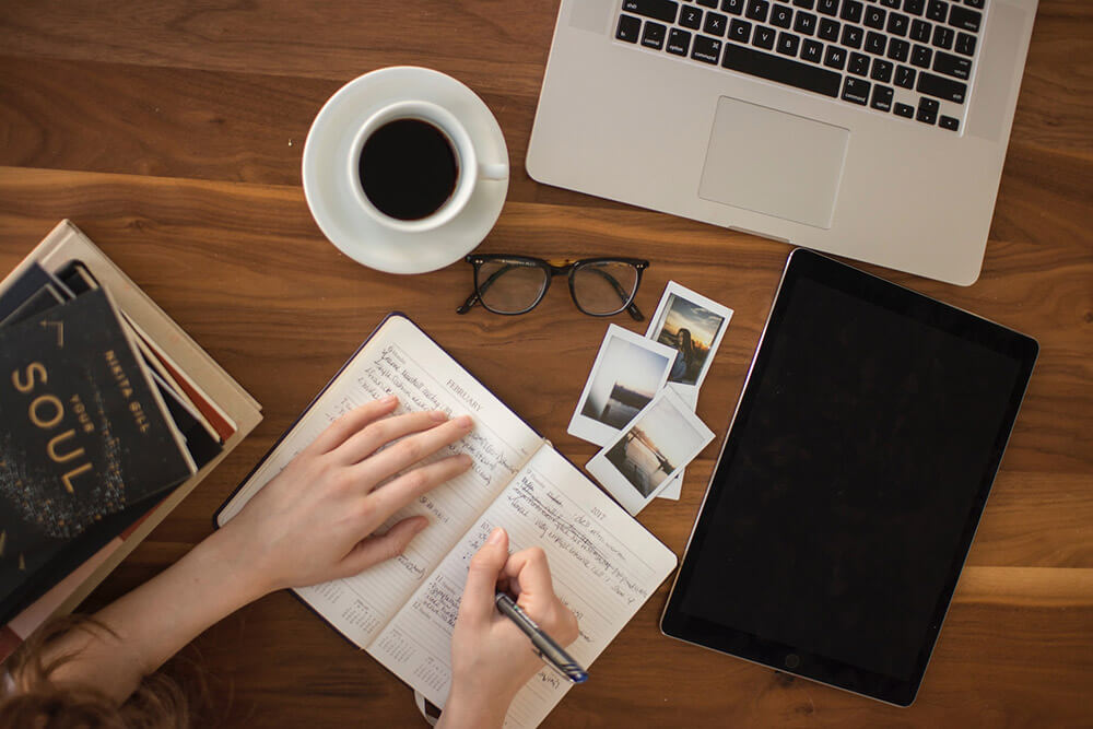 Overhead shot of a desk with books, a laptop, glasses, coffee, photos, and someone writing in a notebook.